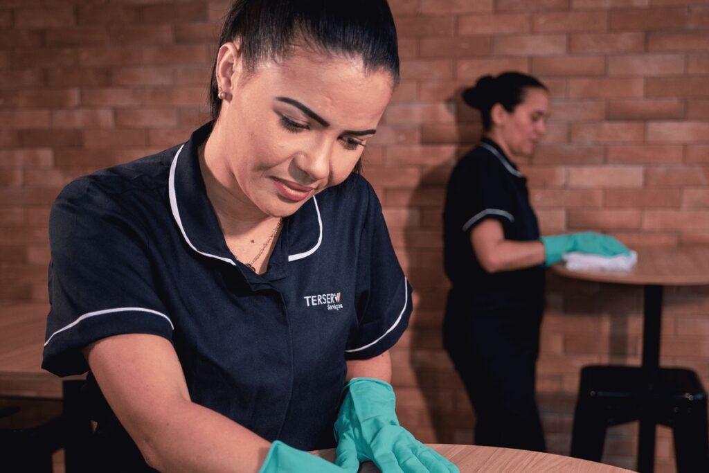 Profissional de limpeza da Terserv, de uniforme, higienizando uma mesa; ao fundo, outra colaboradora trabalhando.